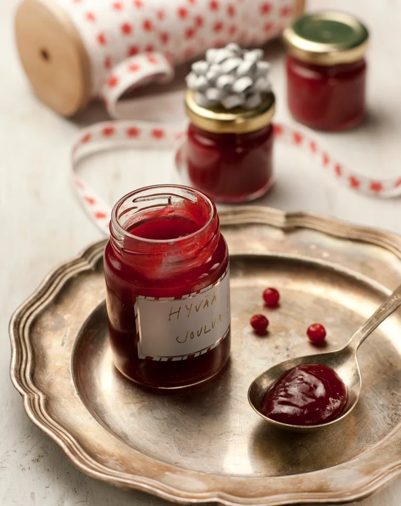 Puolukkaista sinappia lusikassa ja purkissa hopeavadilla, taustalla lahjapurkeissa sinappia ja lahjanauhaa